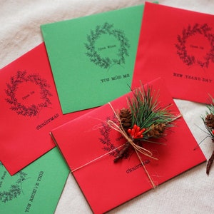 May include: Five red and green envelopes with a wreath design and text. The text on the envelopes reads "Open When", "Christmas", "You Miss Me", "New Year's Day", and "You Receive This".