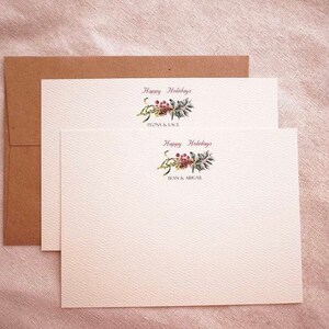 May include: Two white cards with a brown envelope. The cards have a watercolor illustration of a green and red floral sprig with the text "Happy Holidays" and the names "Penny & Cole" and "Jean & Ariel" printed below.