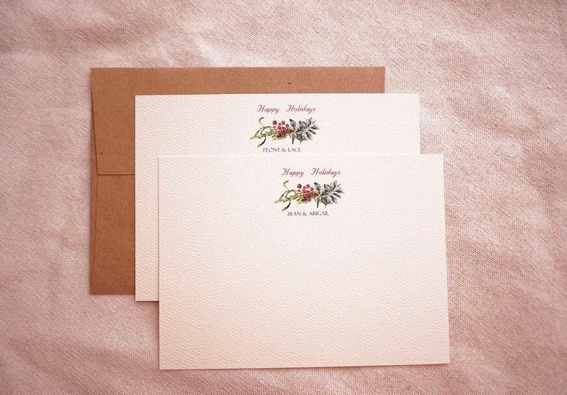 May include: Two white cards with a brown envelope. The cards have a watercolor illustration of a green and red floral sprig with the text "Happy Holidays" and the names "Penny & Cole" and "Jean & Ariel" printed below.