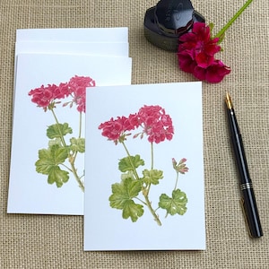 May include: White greeting cards featuring a watercolor illustration of red geranium flowers with green leaves. A black fountain pen, a bottle of ink, and a red flower are also present.