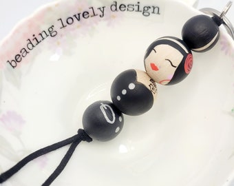 May include: Black and white beaded keychain with a painted face and red lips. The keychain has a black cord and the words "beading lovely design" are written on a white surface behind the keychain.