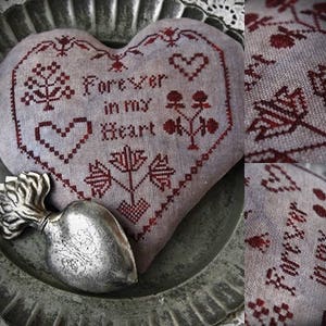 May include: A heart-shaped cushion embroidered with the phrase "Forever in my Heart" in burgundy thread. The design includes floral and heart patterns. A silver heart-shaped ornament sits on a textured metal surface.