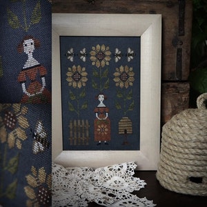 May include: A cross-stitch embroidery design featuring a woman standing in a field of sunflowers. The woman is wearing a red dress and holding a sunflower. The design is framed in a white frame.