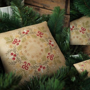 Könnte beinhalten: Ein beiges Kreuzstichkissen mit einem Weihnachtskranz-Design. Der Kranz hat rote Schleifen, Beeren, grüne Tannennadeln und gelbe Sterne. Das Kissen ist von grünen Tannenzweigen umgeben.