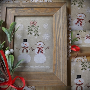 Può includere: Un ricamo a punto croce incorniciato con due pupazzi di neve in piedi sotto un ramo di vischio. I pupazzi di neve indossano cappelli neri e sciarpe rosse. Lo sfondo è un tessuto color crema con fiocchi di neve e un bordo bianco.