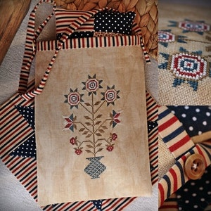 May include: A beige canvas tote bag with red, white, and blue striped trim. The bag features a cross-stitch design of a vase with flowers in red, white, and blue. The bag is perfect for carrying everyday essentials.