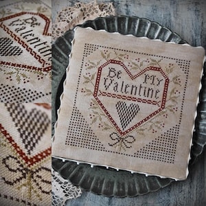 Puede incluir: Un sampler de punto de cruz beige con un corazón rojo y el texto "Be My Valentine". El corazón está rodeado de hojas marrones y verdes y un lazo marrón.