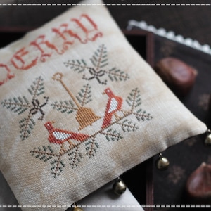 Puede incluir: Una almohada beige con un diseño de punto de cruz que presenta dos pájaros rojos en una rama con hojas verdes. La almohada tiene la palabra "Merry" bordada en hilo rojo en la parte superior y tiene pequeñas campanas unidas a las esquinas inferiores.