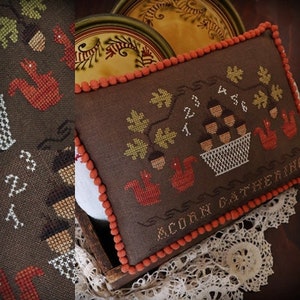 Könnte beinhalten: Ein braunes Kissen mit einem Kreuzstich-Design eines Korbes mit Eicheln, Blättern und Eichhörnchen. Das Kissen hat einen roten und weißen Besatz und der Text "Acorn Gathering" ist unten gestickt.