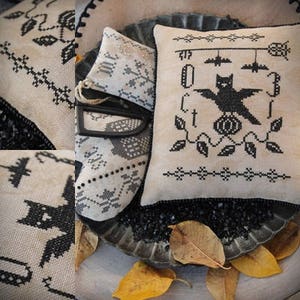Puede incluir: Una colección de artículos de punto de cruz con temática de Halloween. Incluye un cojín con un diseño de murciélago negro, un cojín pequeño con un patrón de copo de nieve y una plancha negra. Los diseños están bordados con hilo negro sobre tela color crema. La imagen también incluye hojas secas.