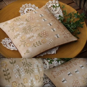 May include: A beige pillow with a cross-stitch design featuring a variety of flowers and bees. The pillow is embroidered with the alphabet in a cursive font. The pillow is sitting on a crocheted doily.