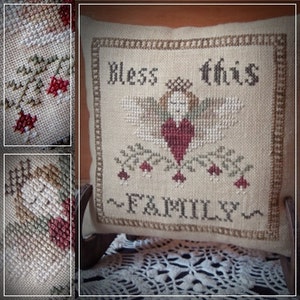 May include: A beige pillow with a cross-stitch design featuring an angel with wings, a heart, and the text "Bless this Family".