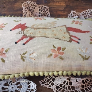 May include: A beige pillow with a green pom-pom trim features an embroidered fox with wings and a floral pattern. The fox is wearing a yellow dress with white polka dots.