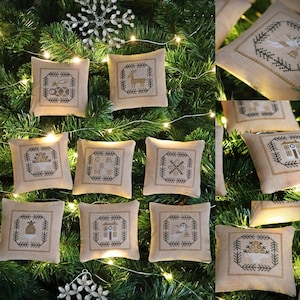 May include: Twelve beige fabric ornaments with embroidered Christmas designs, including a snowflake, a house, a reindeer, a Christmas tree, and a bell. The ornaments are hanging on a green Christmas tree with white fairy lights.