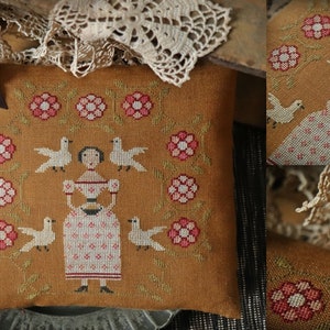 Könnte beinhalten: Senffarbenes quadratisches Kissen mit Kreuzstichmuster. Das Design zeigt eine Frau, die einen Korb hält, umgeben von weißen Tauben und rosa Blumen. Eine schwarze Schleife ist befestigt. Wahrscheinlich ein Dekorationsartikel.