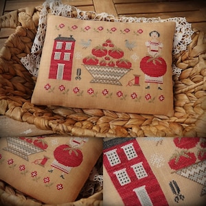 Puede incluir: Una almohada de punto de cruz con una casa roja, una cesta de tomates rojos y una mujer sosteniendo un tomate. La almohada está sobre una cesta tejida.