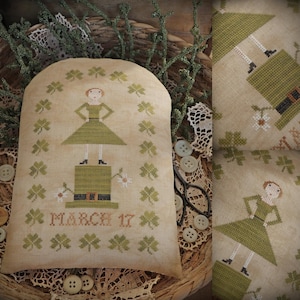 Puede incluir: Un cojín beige bordado a punto de cruz con una figura vestida de verde de pie sobre un sombrero de duende verde. El cojín está decorado con tréboles verdes y el texto "MARCH 17". El cojín está rodeado de elementos decorativos.