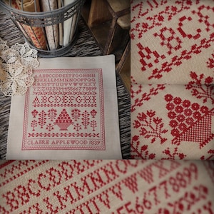 Könnte beinhalten: Ein Kreuzstich-Sampler mit rotem Faden auf cremefarbenem Stoff. Das Design umfasst das Alphabet, Zahlen und florale Motive. Der Text "CLAIRE APPLEWOOD 1839" ist unten gestickt. Der Sampler wird mit anderen Vintage-Artikeln präsentiert.