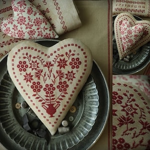 Op de afbeelding: Drie hartvormige geborduurde kussens met rode en witte bloemenpatronen. De kussens liggen op een rustieke achtergrond met een metalen dienblad.