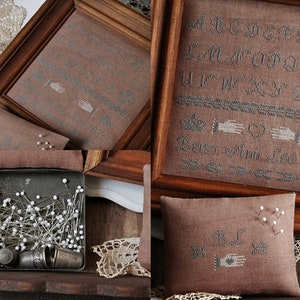 Könnte beinhalten: Eine Sammlung von Kreuzstich-Samplern und Zubehör im Vintage-Stil. Das Bild zeigt gerahmte Sampler mit gestickten Alphabeten und Motiven, zusammen mit einem kleinen Kissen und einer Metallbox voller Nadeln und Fingerhüte. Die Farbpalette ist gedämpft, mit Braun- und Grautönen.