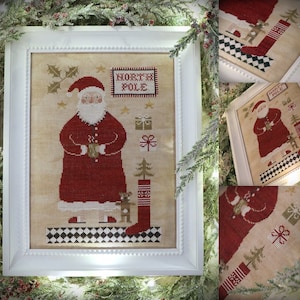 May include: A cross-stitch embroidery of Santa Claus holding a mug, standing in front of a stocking and a Christmas tree. The embroidery is framed in white and has the text "North Pole" above Santa.