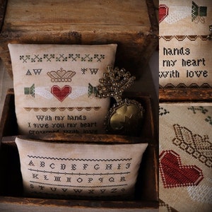 Op de afbeelding: Een kruissteek-sampler met een hart en de tekst "With my hands I give you my heart crowned with love". De sampler wordt getoond in een houten doos met een decoratief metalen ornament. Het alfabet is ook op een aparte sampler geborduurd.