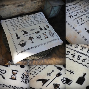 Op de afbeelding: Een vierkant gebroken wit stoffen kussen met zwarte kruissteek borduursels. Het ontwerp bevat het alfabet, de cijfers nul tot en met negen en iconen zoals een heksenhoed, bezem, ketel, zandloper, sleutel, slang en flessen.