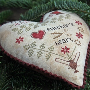 May include: A heart-shaped pillow with a cross-stitch design featuring a red heart, green leaves, and the text "Stitcher's Heart". The pillow has a burgundy trim.
