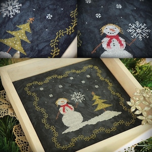 Peut inclure: Une broderie au point de croix sur tissu sombre représentant un bonhomme de neige blanc avec une écharpe rouge tenant un flocon, un sapin doré et des flocons blancs, encadrée par une bordure de vigne dorée dans un cadre carré en bois clair.