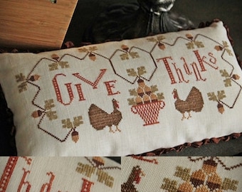 May include: Rectangular, off-white pillow with cross-stitched design. The words "GIVE THANKS" are stitched in red, with images of turkeys, a basket of fruit, and autumn leaves. The pillow has a ruffled, brown border.