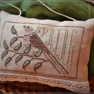 Op de afbeelding: Een beige kussen met een witte kantversiering heeft een kruissteekontwerp van een vogel die op een tak zit met groene bladeren. Het ontwerp bevat het woord "Lente" in een gestileerd lettertype.