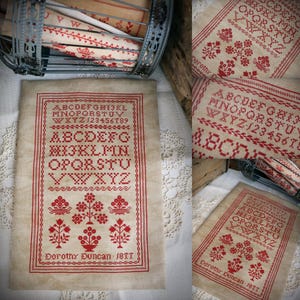 Peut inclure: Un point de croix de style vintage avec du fil rouge sur un tissu crème. Le motif comprend l'alphabet, les chiffres et des motifs floraux. Le texte "Dorothy Duncan 1877" est en bas.