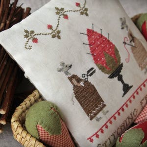 May include: A white pillow with a cross-stitch design featuring two mice, a strawberry, and the text "Stitching Fruit." The pillow is sitting on a woven basket with a green and red fabric strawberry.