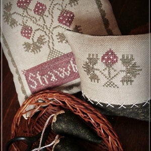 Puede incluir: Dos fundas de cojín bordadas con un diseño floral. La primera funda de cojín presenta un diseño floral verde y marrón con fresas rojas y el texto "Straw". La segunda funda de cojín presenta un diseño floral verde con una sola fresa roja. Ambas fundas de cojín están sobre una superficie de madera marrón.