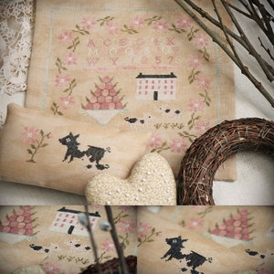 May include: A cross-stitch sampler featuring a house, flowers, and a cow. The sampler is framed and has a light brown background. The text on the sampler reads "ABCDEFGHIJKLMNOPQRSTUVWXYZ 12345678".
