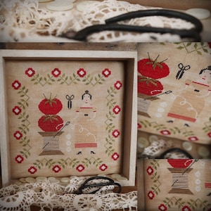 Peut inclure: Une broderie au point de croix encadrée représentant deux femmes cousant avec une bobine de fil et des tomates rouges. La broderie est entourée d'une vigne verte avec des fleurs rouges.