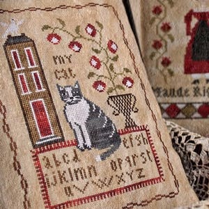 Könnte beinhalten: Ein beigefarbenes Kissen mit einem Kreuzstich-Design, das eine Katze, ein Haus, eine Pflanze und das Alphabet zeigt. Das Kissen liegt auf einer braunen Oberfläche mit einer weißen Spitzenüberlage.