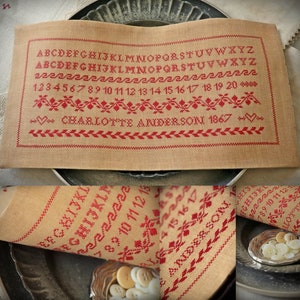 Può includere: Un primo piano di un sampler in lino beige con ricamo rosso. Il sampler presenta l'alfabeto, i numeri da 1 a 20, un bordo floreale e il testo "CHARLOTTE ANDERSON 1867".