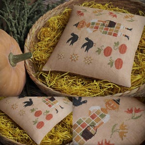 May include: A beige cushion with a cross-stitch design of a scarecrow, pumpkins, and crows. The cushion is in a woven basket with yellow straw, and a pumpkin is visible. The autumn-themed pattern is detailed and colourful.