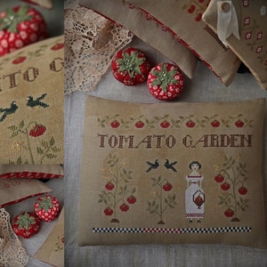 Op de afbeelding: Een beige kussen met een kruissteekontwerp van een tomaten tuin. Het ontwerp toont een meisje dat een mandje vasthoudt, twee vogels en tomatenplanten met rode tomaten. Het kussen heeft de tekst "TOMATO GARDEN" geborduurd in bruin garen.