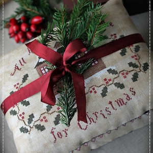 Può includere: Un cuscino bianco con un nastro rosso legato intorno. Il cuscino è ricamato con le parole "All for Christmas" e un piccolo disegno di una casa. Un rametto di pino verde è posto sopra il nastro.