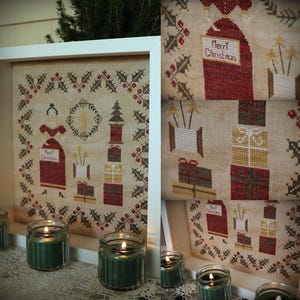 Könnte beinhalten: Kreuzstich-Weihnachtssampler in einem weißen Rahmen, mit einer Frau in rotem Kleid, einem Weihnachtsbaum und gestapelten Geschenken. Der Sampler enthält den Text "Merry Christmas" und ist von einem Stechpalmen- und Beerenrand umgeben. Grüne Kerzen in Gläsern sind im Vordergrund.