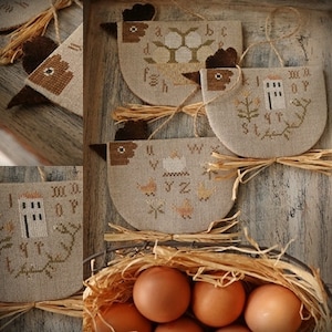 Puede incluir: Cuatro adornos de punto de cruz marrones y blancos con forma de pollos con diferentes letras del alfabeto en cada uno. Los adornos cuelgan de una superficie de madera rústica. Una cesta de huevos marrones se encuentra en la esquina inferior derecha de la imagen.