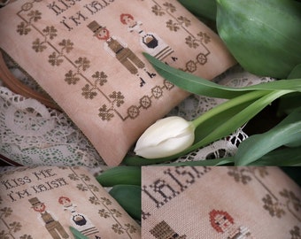 May include: A cross-stitched pillow with the text "Kiss Me I'm Irish" and images of an Irish couple. The pillow is beige with brown stitching and a border of shamrocks. A white tulip rests nearby.