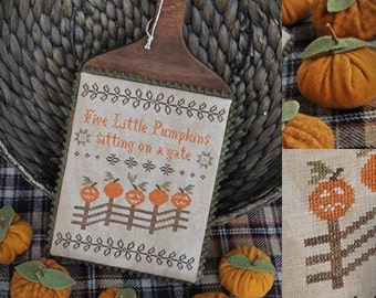 May include: A cross-stitch pattern on a wooden plaque with the text "Five Little Pumpkins Sitting on a Gate". The pattern features five pumpkins with faces, a fence, and a gate. The plaque is surrounded by small orange pumpkins.