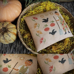 Könnte beinhalten: Ein beiges quadratisches Kissen mit einem Kreuzstichdesign mit einer Vogelscheuche, schwarzen Vögeln, Kürbissen und Blumen. Das Kissen befindet sich in einem geflochtenen Korb mit gelbem Stroh, mit zwei Kürbissen im Hintergrund.
