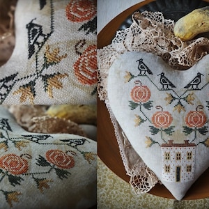 Puede incluir: Una almohada en forma de corazón con un diseño de punto de cruz que presenta una pequeña casa, calabazas y pájaros. La almohada está sobre una superficie de madera marrón con un encaje blanco.