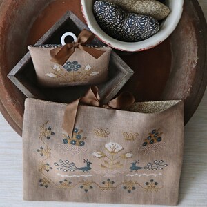 Könnte beinhalten: Zwei braune Leinentaschen mit gestickten Blumen- und Vogelmotiven. Die kleinere Tasche hat ein braunes Band umgeschlungen. Die größere Tasche hat ein braunes Band umgeschlungen und liegt auf einer Holzoberfläche.