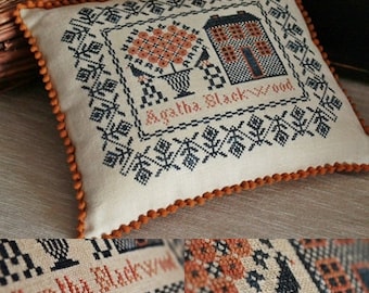 May include: A white pillow with a brown trim features a cross-stitch design of a house, a tree, and the text "Agatha Blackwood".