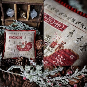May include: A beige and brown cross-stitch pillow with a red sleigh carrying presents and a teddy bear. The pillow has the text "HO-HO-HO" stitched on it. The pillow is surrounded by pine cones and evergreen branches.